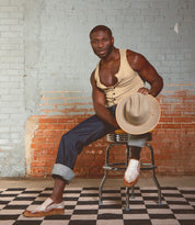 A man embodies casual sophistication in a beige sleeveless shirt and jeans, seated on a stool with a hat in hand. Against a brick wall and checkerboard floor, his look is finished with Bed Stu Freestyle sneakers featuring subtle leather detailing.