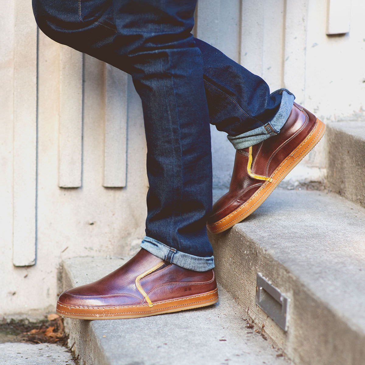 Sporting Freestyle shoes from Bed Stu, a person in jeans stands on concrete steps, exuding casual sophistication with their brown leather footwear accented by cream stitching.