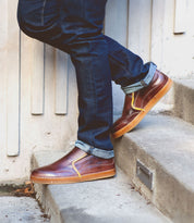 Sporting Freestyle shoes from Bed Stu, a person in jeans stands on concrete steps, exuding casual sophistication with their brown leather footwear accented by cream stitching.