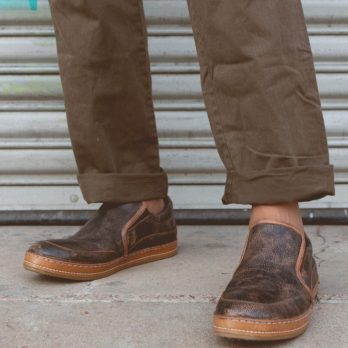 A person wearing Bed Stu's Freestyle slip-on shoes in dark brown, featuring subtle leather detailing, and olive green pants stands near a metallic garage door, exuding casual sophistication.