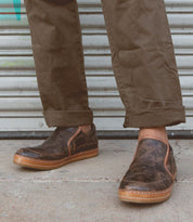 A person wearing Bed Stu's Freestyle slip-on shoes in dark brown, featuring subtle leather detailing, and olive green pants stands near a metallic garage door, exuding casual sophistication.