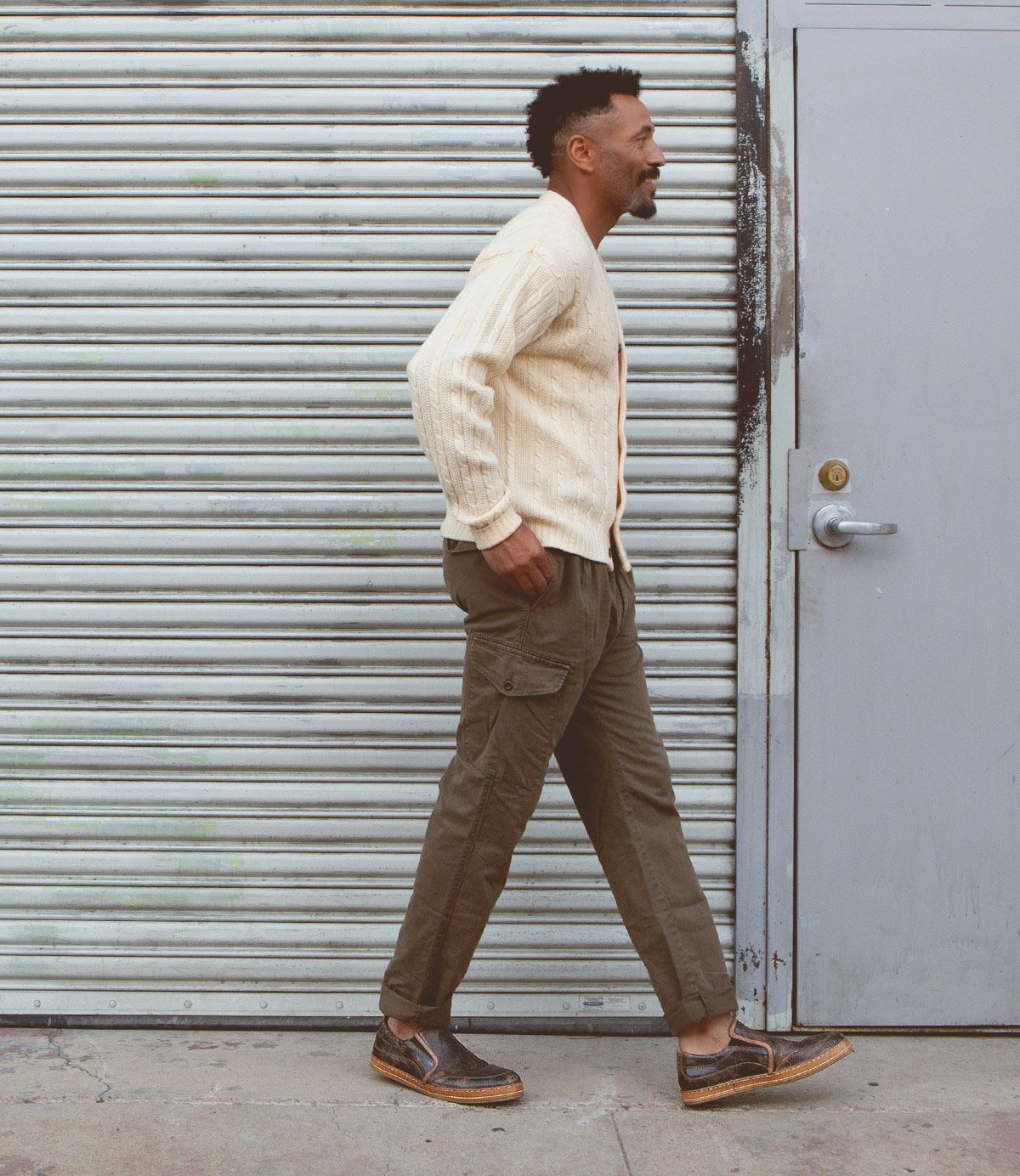 A man with a short haircut wearing a cream cardigan, olive cargo pants, and Bed Stu's Freestyle shoes in brown with leather detailing walks past a corrugated metal shutter and a gray door, exuding casual sophistication.