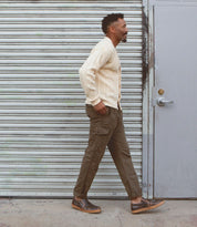 A man with a short haircut wearing a cream cardigan, olive cargo pants, and Bed Stu's Freestyle shoes in brown with leather detailing walks past a corrugated metal shutter and a gray door, exuding casual sophistication.