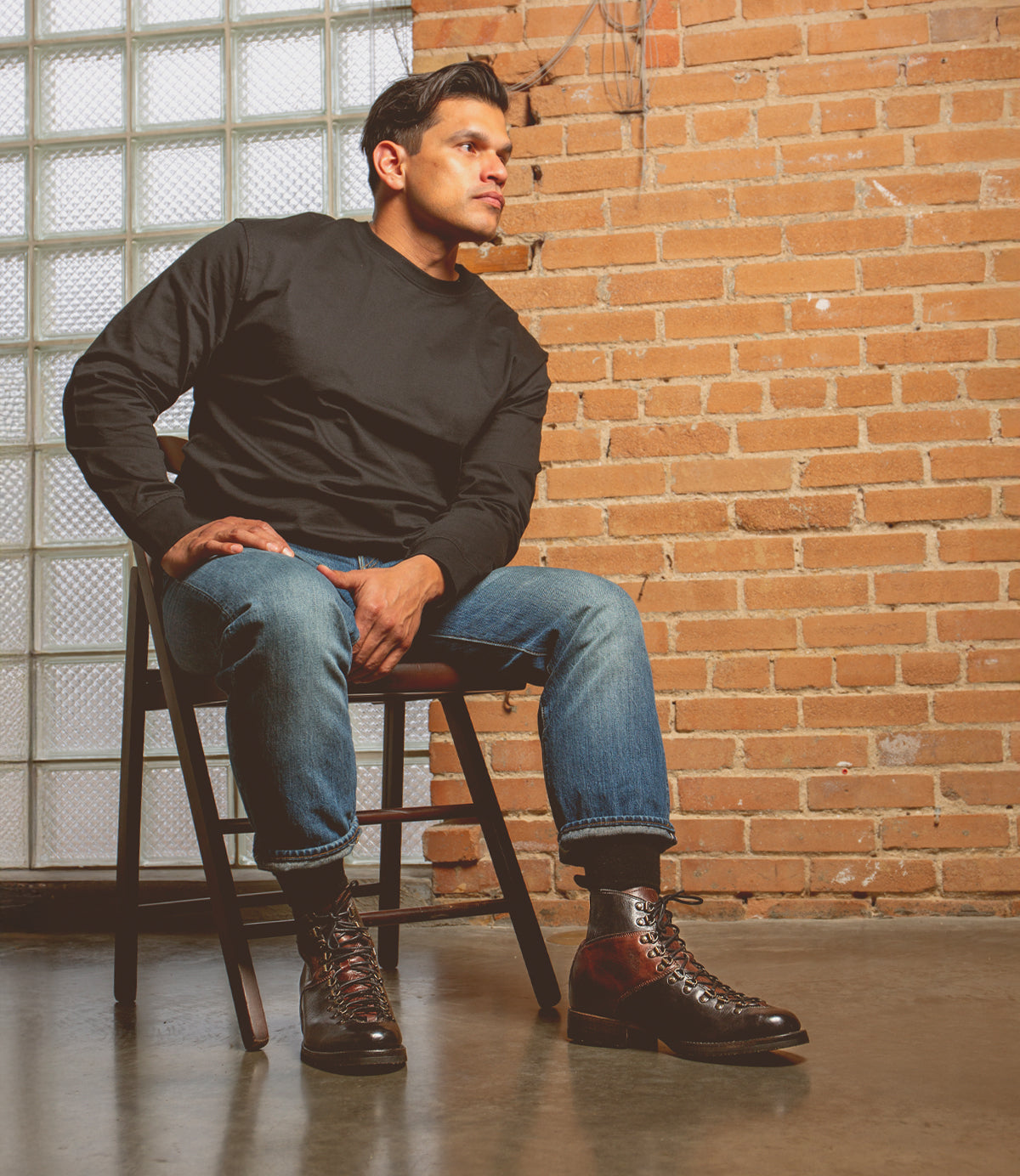 Seated on a wooden chair sporting Bed Stu's Barton high-top sneakers in leather, a black long-sleeve shirt, and blue jeans, this person completes their stylish outfit. The comfort of breathable insoles adds to the chic look in front of a brick wall with a glass block window.