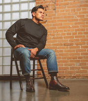 Seated on a wooden chair sporting Bed Stu's Barton high-top sneakers in leather, a black long-sleeve shirt, and blue jeans, this person completes their stylish outfit. The comfort of breathable insoles adds to the chic look in front of a brick wall with a glass block window.