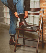 A person in jeans and a black shirt is tying the laces of their Bed Stu Barton brown leather boots, which feature a breathable insole, while resting one foot on a wooden chair.