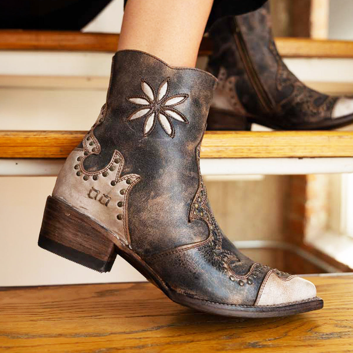 A person wearing the Bed|Stü Frio distressed leather Western bootie, featuring floral embroidery and antiqued brass studs, sits on a wooden step.