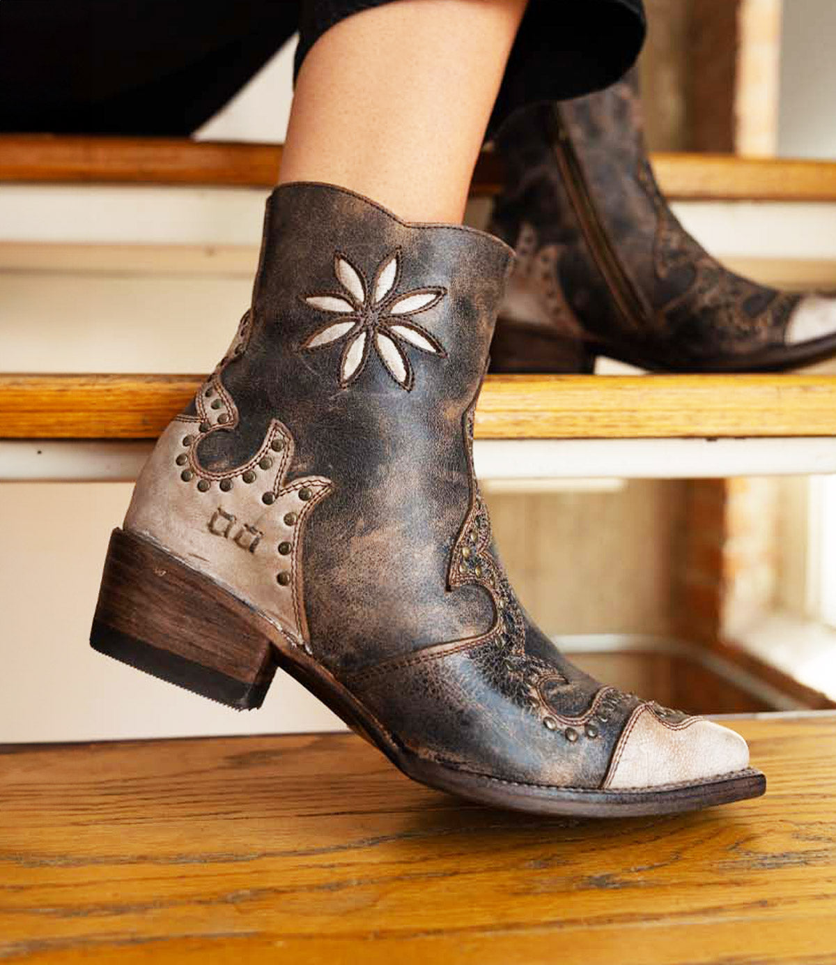 A person wearing the Bed|Stü Frio distressed leather Western bootie, featuring floral embroidery and antiqued brass studs, sits on a wooden step.