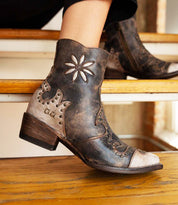 A person wearing the Bed|Stü Frio distressed leather Western bootie, featuring floral embroidery and antiqued brass studs, sits on a wooden step.