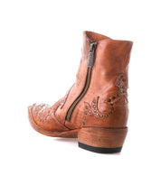 The Bed Stu Frio is a brown leather Western bootie featuring brass studs, floral cutout details, a side zipper, and a low stacked heel, shown on a white background.