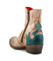 The Bed Stu Frio is a tan and teal leather Western bootie featuring a side zipper, decorative stitching, brass studs, floral cutout accents, and a low wooden heel, displayed from a rear side angle.