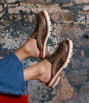 A person wears Bed Stu Fuel brown handwoven leather slip-on loafers with thick white, lightweight outsoles and blue jeans, resting their feet against a worn, textured brick wall.