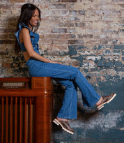A woman in a blue sleeveless top and flared jeans sits sideways on a wooden cabinet against a distressed brick wall, smiling as she looks down. Her Bed Stu Fuel handwoven leather shoes complete her relaxed, stylish vibe.