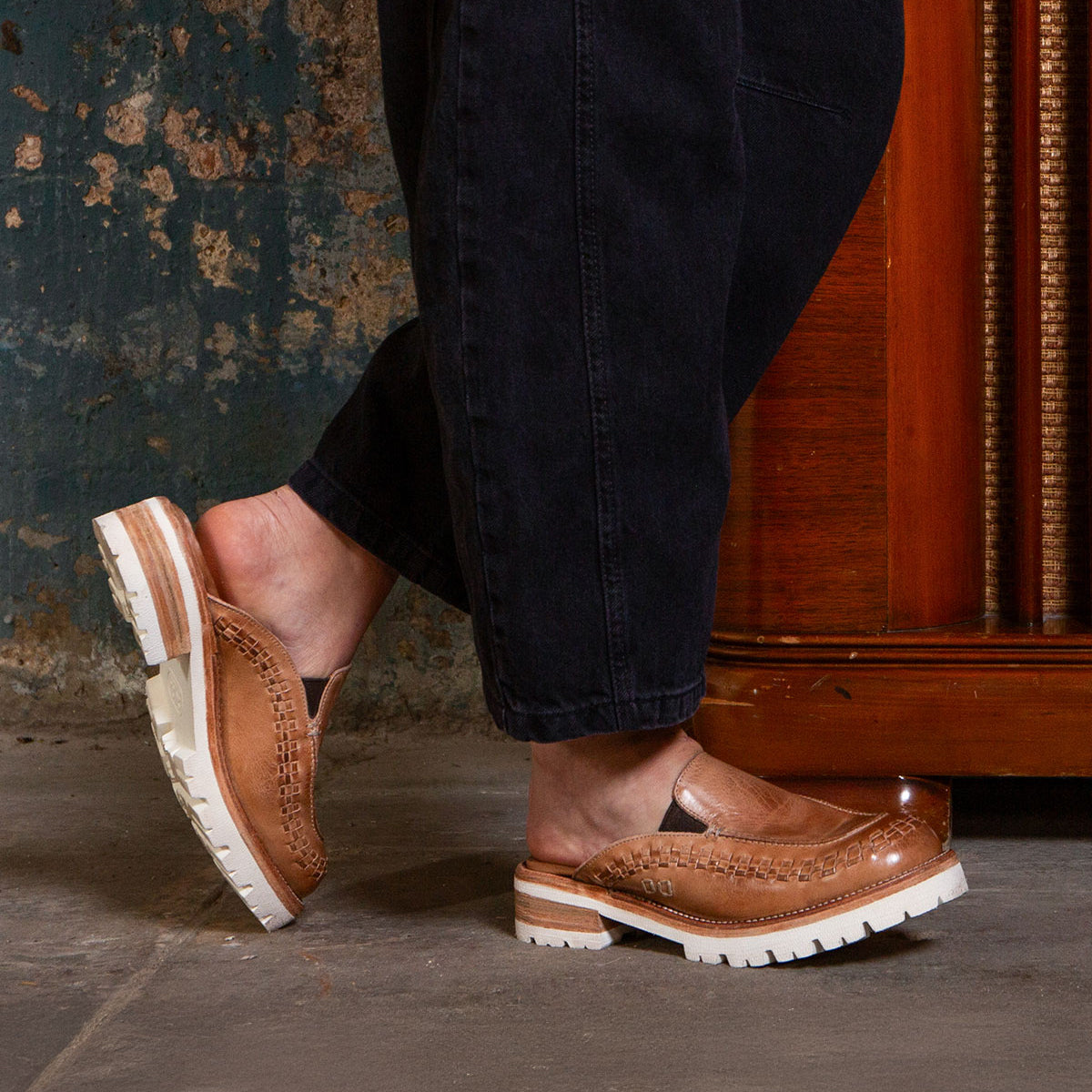 A person stands indoors on a concrete floor, wearing black pants and Bed Stu's Fuel tan handwoven leather slip-on shoes with thick, lightweight outsoles.