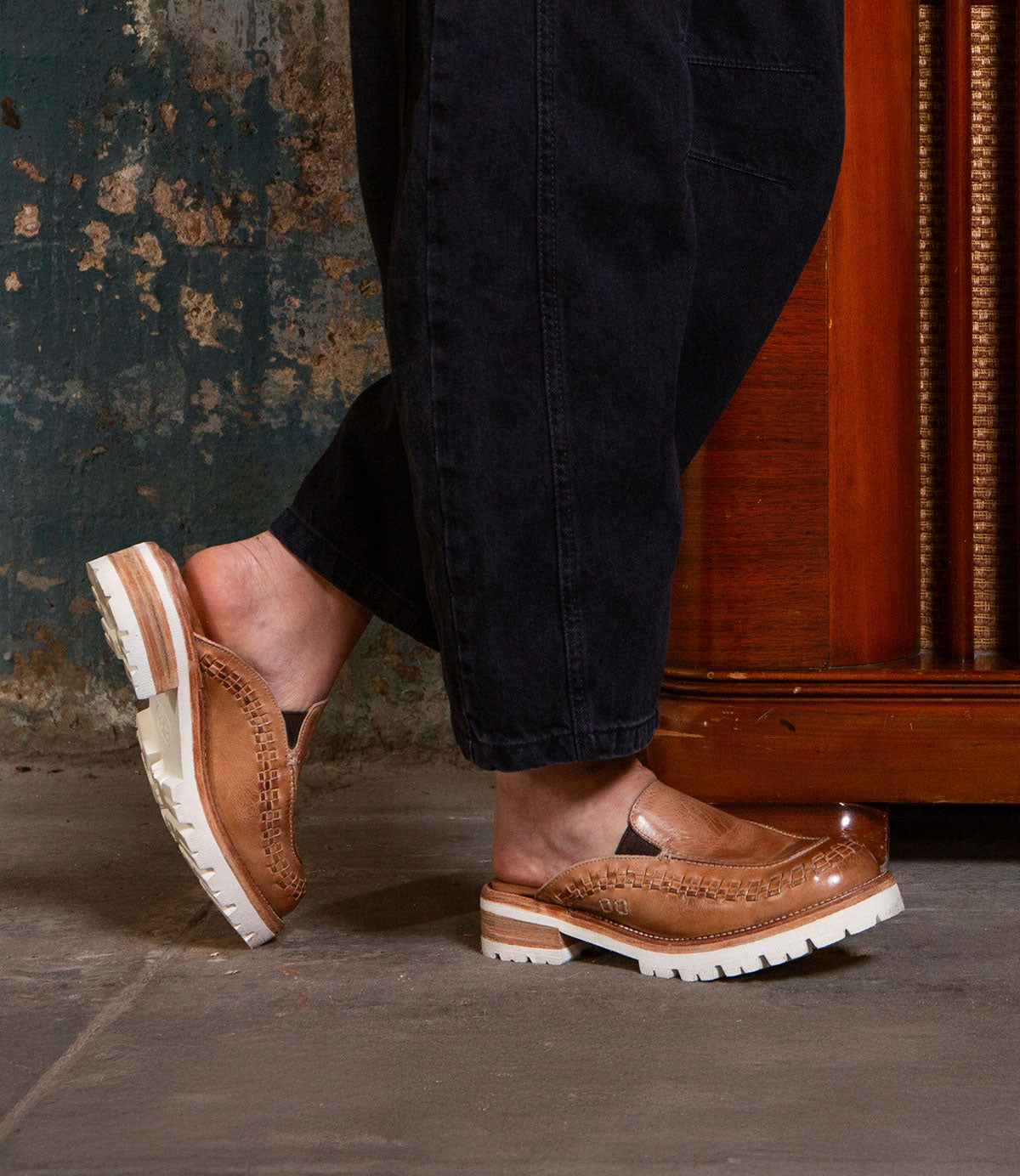 A person stands indoors on a concrete floor, wearing black pants and Bed Stu's Fuel tan handwoven leather slip-on shoes with thick, lightweight outsoles.