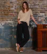 A woman in a beige t-shirt and black pants smiles against a weathered brick wall, one hand on a vintage radio—her look finished with Bed Stu's Fuel shoes, featuring a flexible fit and lightweight outsole.