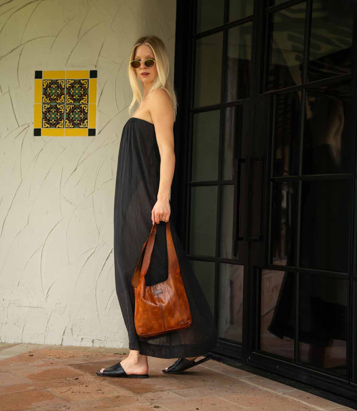 A woman wearing a strapless black dress, sunglasses, and Bed Stu Gailee leather slide sandals with a cushioned footbed stands outside near a glass door, holding a brown bag.