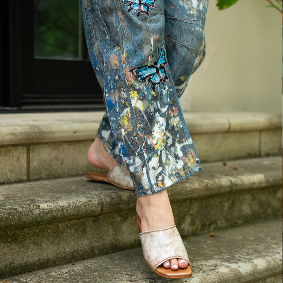 A person in paint-splattered jeans with butterfly patches steps down stone stairs wearing Bed Stu's Gailee leather slides featuring a cushioned footbed.
