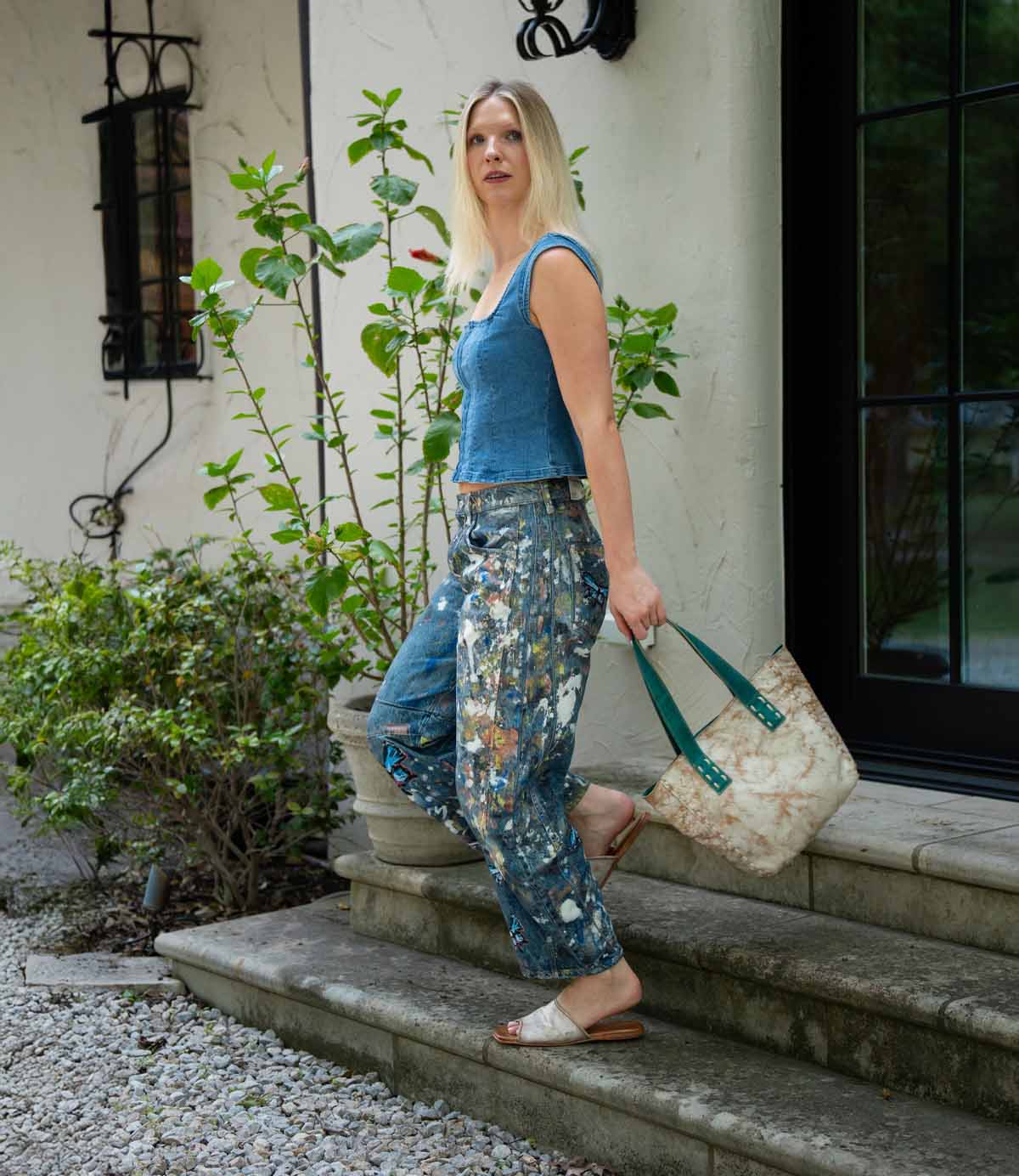 A woman in paint-splattered jeans and a blue sleeveless top stands on stone steps wearing Bed Stu’s Gailee leather slides with cushioned footbed, holding a large beige tote, greenery and a house behind her.