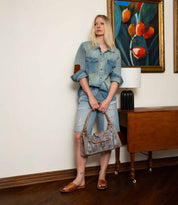 Woman in denim shirt and distressed jean shorts holds a gray handbag indoors by a wooden table and painting, wearing Bed Stu Gailee comfortable brown leather slide sandals with a cushioned footbed.