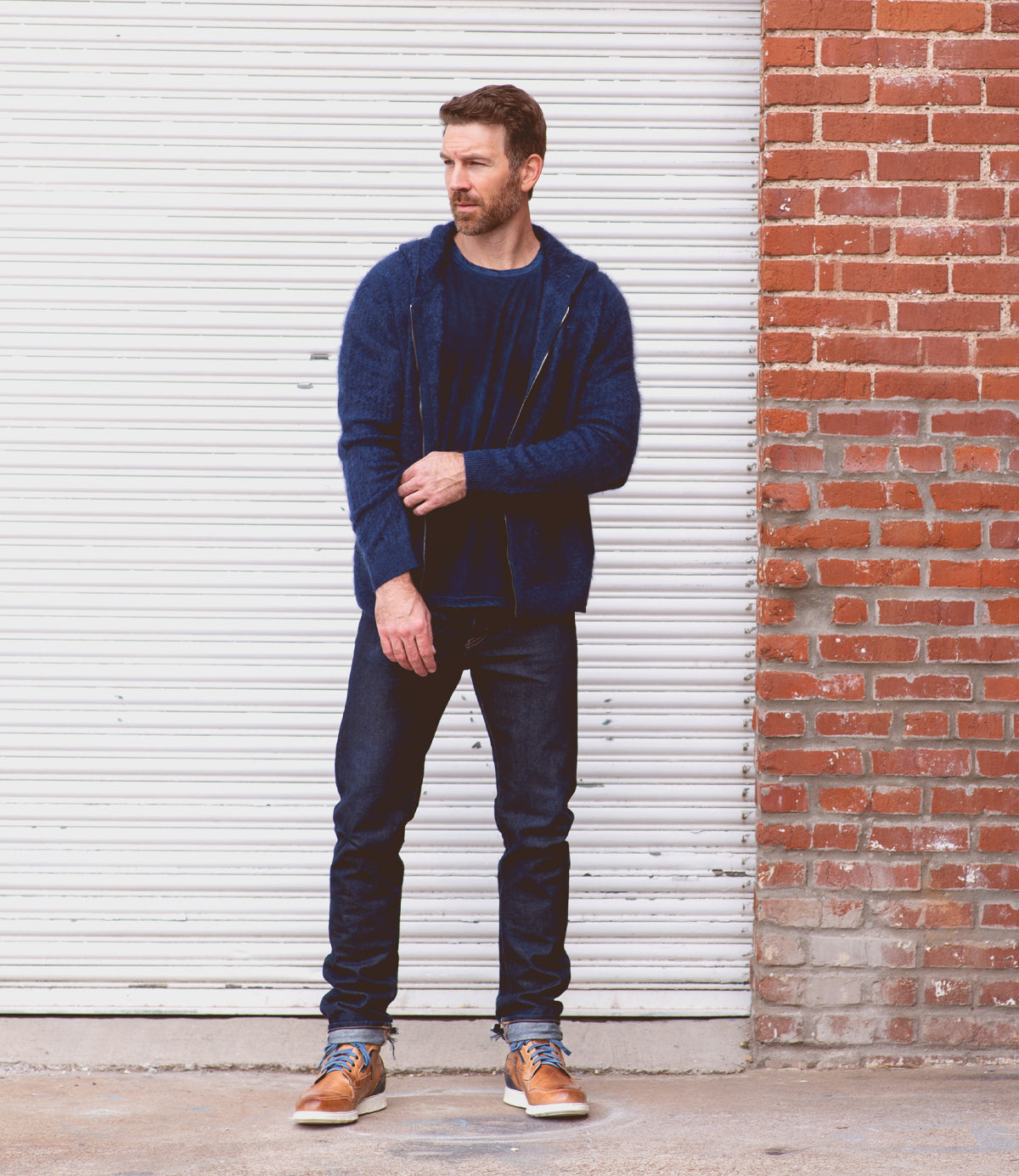 A man in jeans and a hoodie, wearing Bed Stu Galahad leather sneakers, standing in front of a garage.