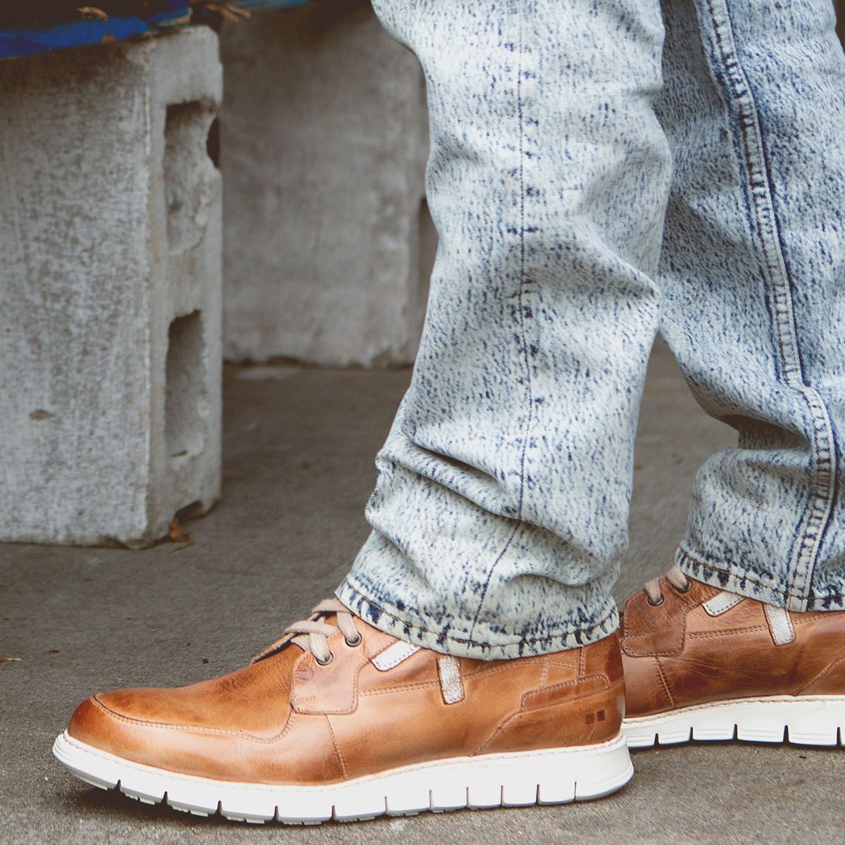 A person wearing light blue jeans and brown Bed Stu Galahad high-top leather sneakers with breathable insoles stands on a concrete surface near some cinder blocks.
