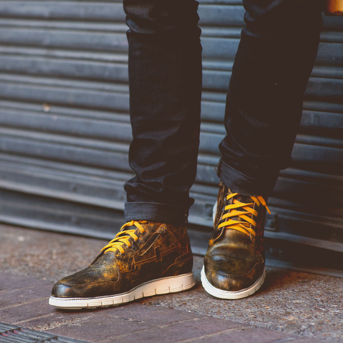 A person wearing the Galahad high-top leather sneakers by Bed Stu, featuring a distressed finish with yellow laces, comfortable insoles, and white soles, stands against a corrugated metal background.