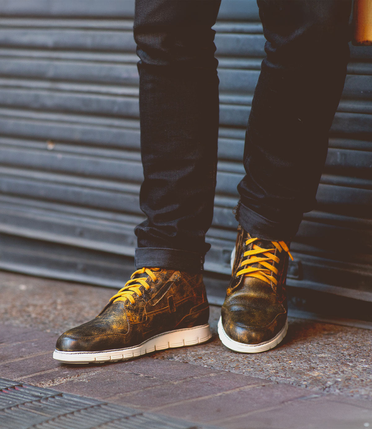 A person wearing the Galahad high-top leather sneakers by Bed Stu, featuring a distressed finish with yellow laces, comfortable insoles, and white soles, stands against a corrugated metal background.