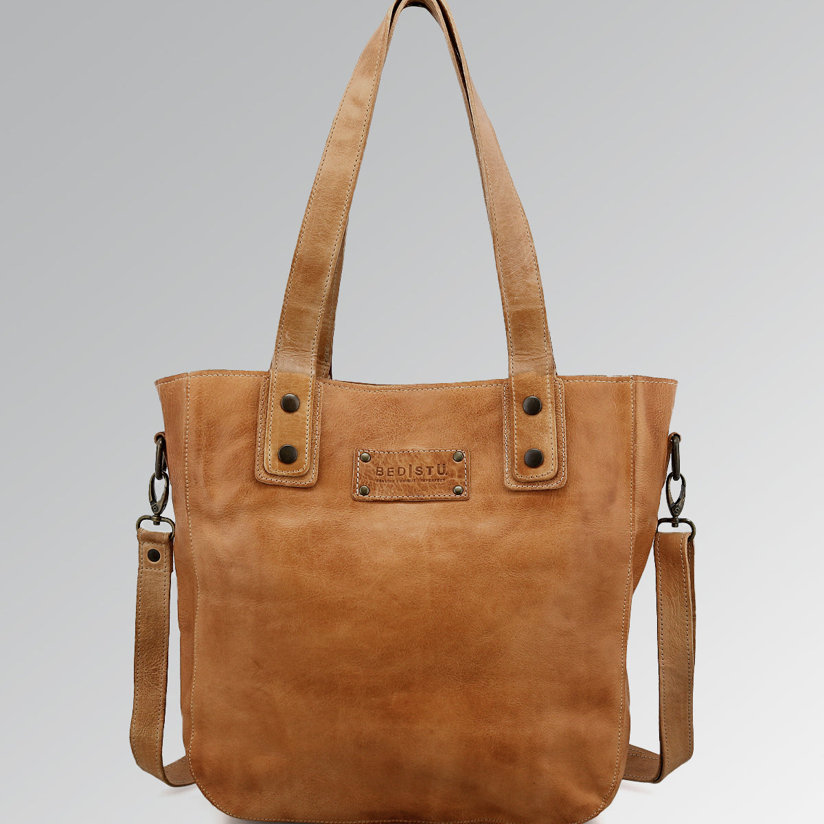 The Bed Stu Studio Gaviota II Body is a tan leather tote featuring dual handles, a detachable shoulder strap, and a stitched front label, shown against a light gray background.