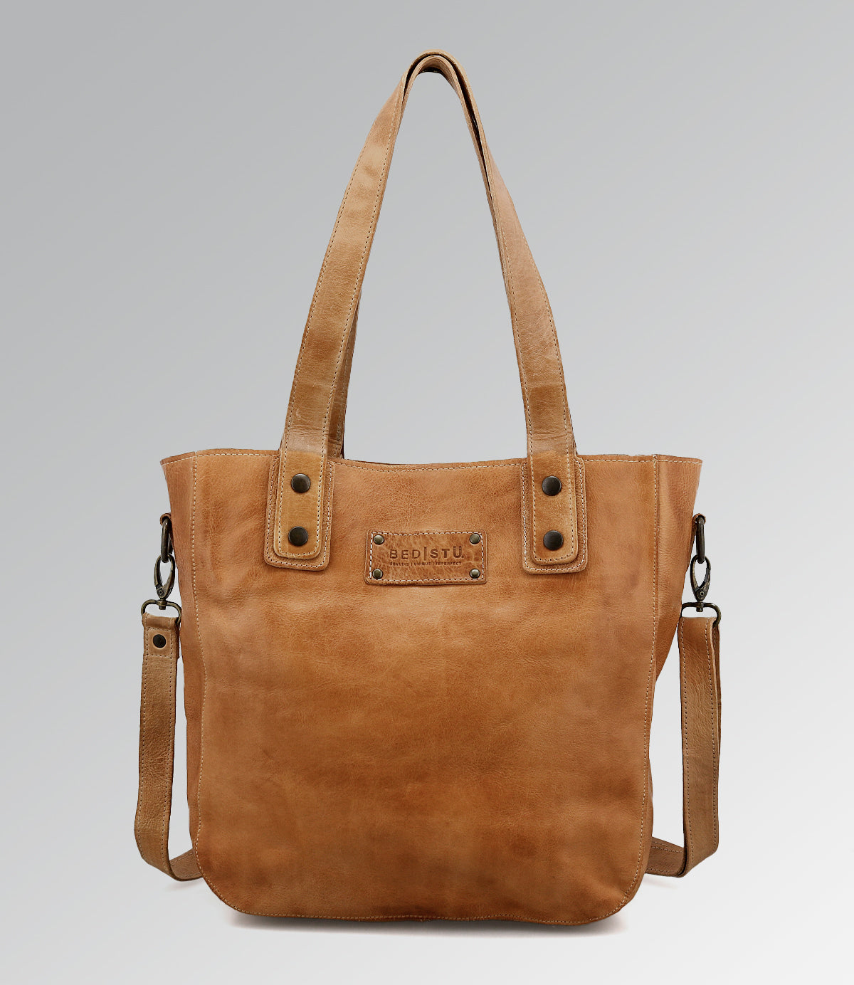 The Bed Stu Studio Gaviota II Body is a tan leather tote featuring dual handles, a detachable shoulder strap, and a stitched front label, shown against a light gray background.