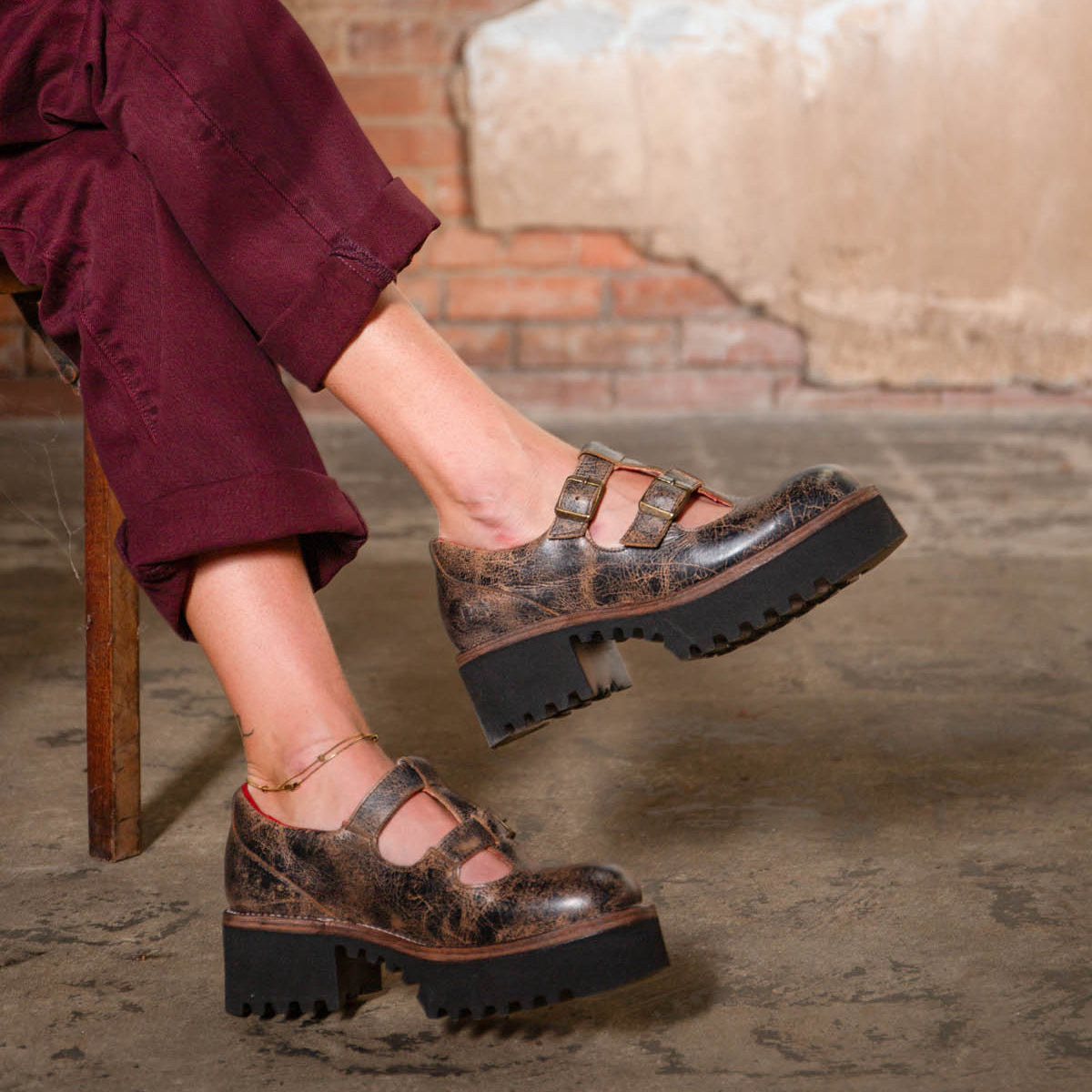 Someone wearing burgundy pants and Bed Stu Gear vintage-style brown women's Mary Jane shoes with a double-strap design and thick black XL Extralight sole sits on a wooden chair.