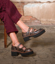 Someone wearing burgundy pants and Bed Stu Gear vintage-style brown women's Mary Jane shoes with a double-strap design and thick black XL Extralight sole sits on a wooden chair.