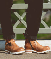 A knee-down shot of a woman in black cuffed denim standing on a brdige with bedStu Ginger II boots in tan on. 