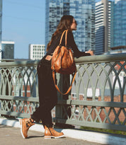 A woman with long hair, sunglasses, and a brown handbag leans against a bridge railing with a cityscape in the background, confidently rocking her Ginger II ankle boots by Bed Stu.