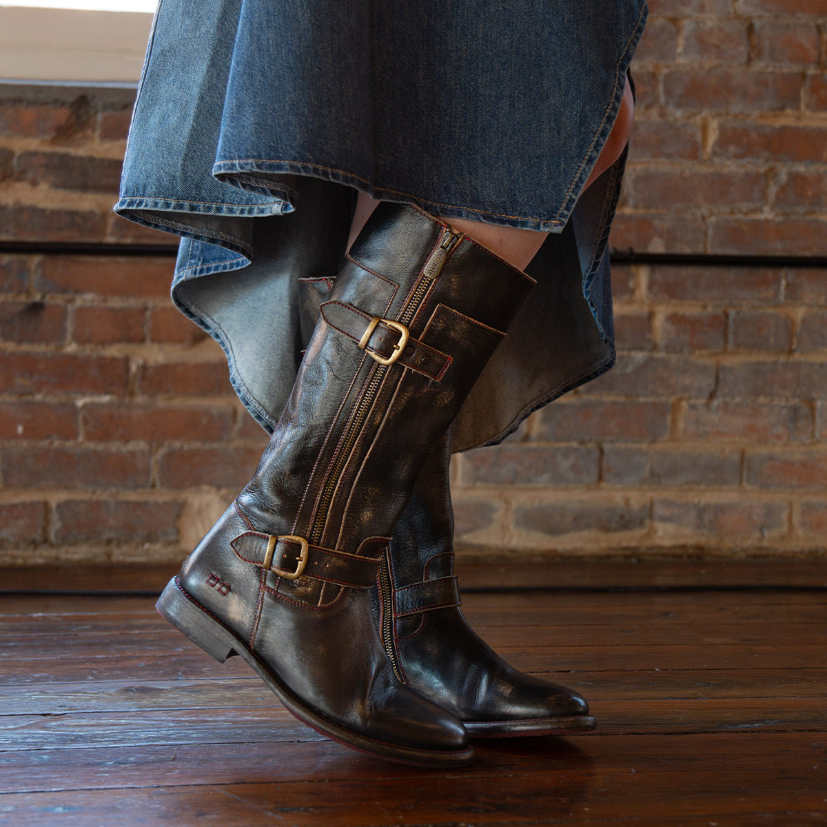 A person wearing a high-low denim skirt and Bed Stu Gogo Lug brown leather moto boots stands on a wooden floor in front of a brick wall.