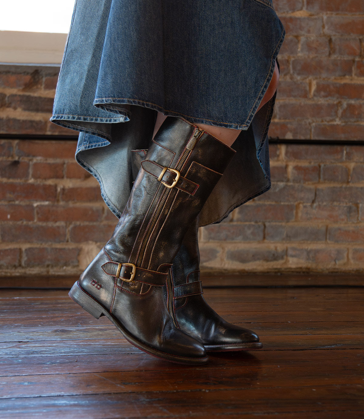 A person wearing a high-low denim skirt and Bed Stu Gogo Lug brown leather moto boots stands on a wooden floor in front of a brick wall.