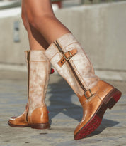 Close-up of a person walking on a paved surface in the Gogo Lug tan leather knee-high boots by Bed Stu, featuring dual zippers, buckle details, and a durable lug outsole.