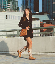 A woman walks confidently on an urban rooftop wearing a black dress, carrying a brown crossbody bag, and sporting Bed Stu's Gogo Lug mid-calf leather boots with durable lug outsoles. City buildings are visible in the background.