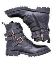 The Bed Stu Grab boots are black leather combat-inspired footwear with studded buckle straps, side zippers, and thick treaded soles—shown with one boot upright and the other on its side.