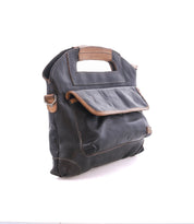 The Greenway by Bed|Stü is a black full-grain leather satchel with brown trim, designed with a front flap, side pocket, rectangular cutout handles, and a convenient zip-top closure. This elegant bag is photographed against a white background.