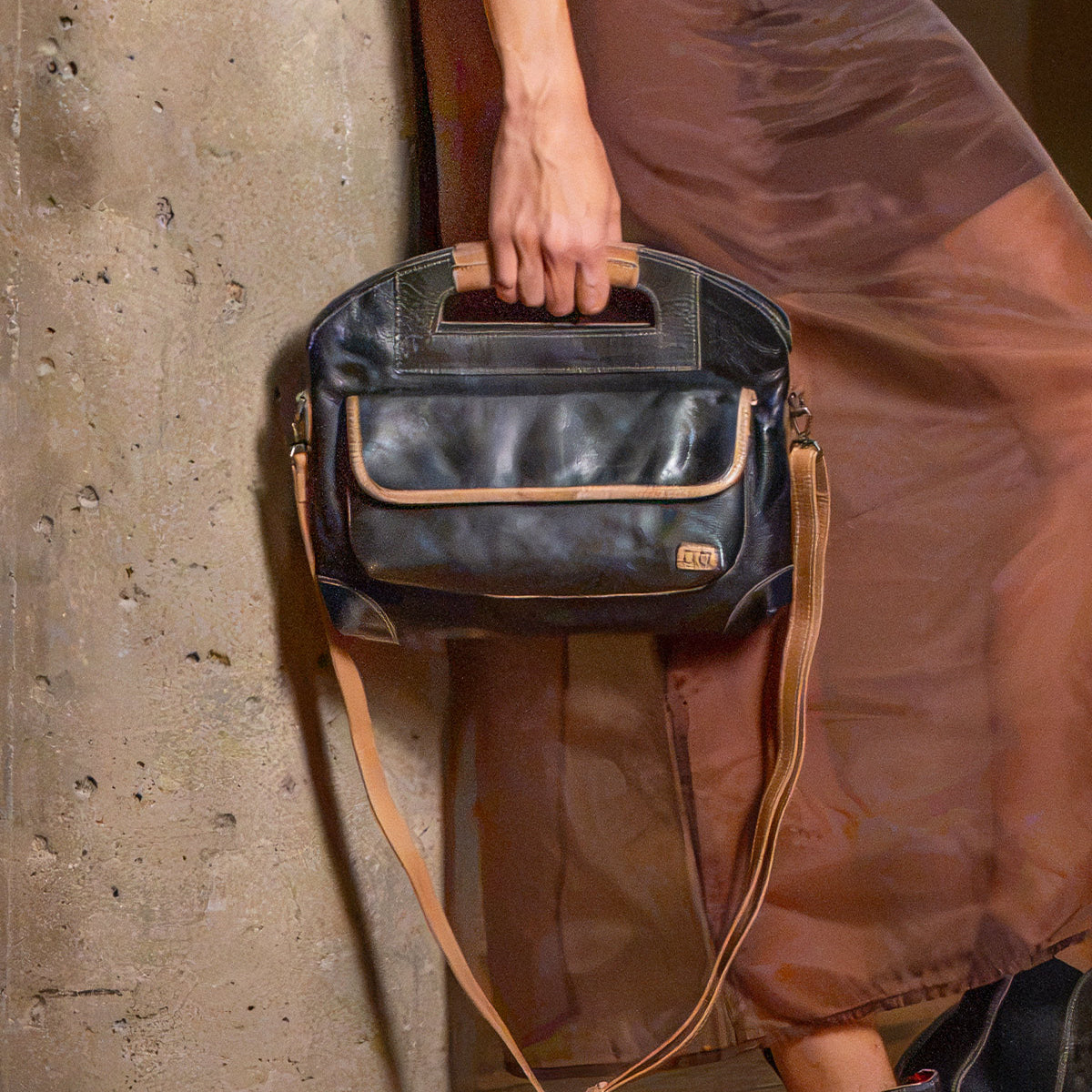 A person in a brown dress holds a two-tone black and tan full-grain leather Greenway handbag featuring a top handle and a detachable crossbody strap.