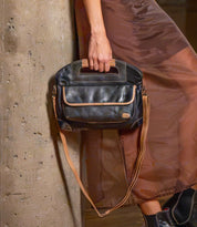 A person in a brown dress holds a two-tone black and tan full-grain leather Greenway handbag featuring a top handle and a detachable crossbody strap.