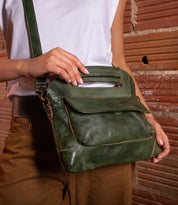 Wearing brown pants and a white top, a person holds the Bed Stu Greenway handbag—with handle, shoulder strap, and zipper—while standing by a brick wall.