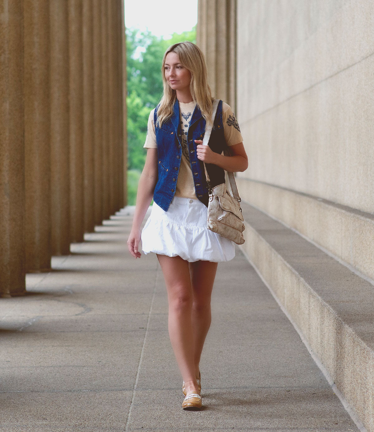 A woman with blonde hair walks along a building with tall columns, wearing a beige top, blue vest, short white skirt, tan shoes, and a Bed Stu Greenway crossbody bag with inside zippered pockets.