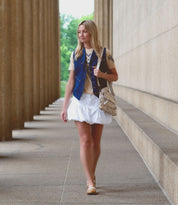 A woman with blonde hair walks along a building with tall columns, wearing a beige top, blue vest, short white skirt, tan shoes, and a Bed Stu Greenway crossbody bag with inside zippered pockets.