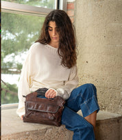 A woman in a cream sweater and blue jeans sits by a window, holding a Bed|Stü Greenway full-grain leather handbag with a zip-top closure.