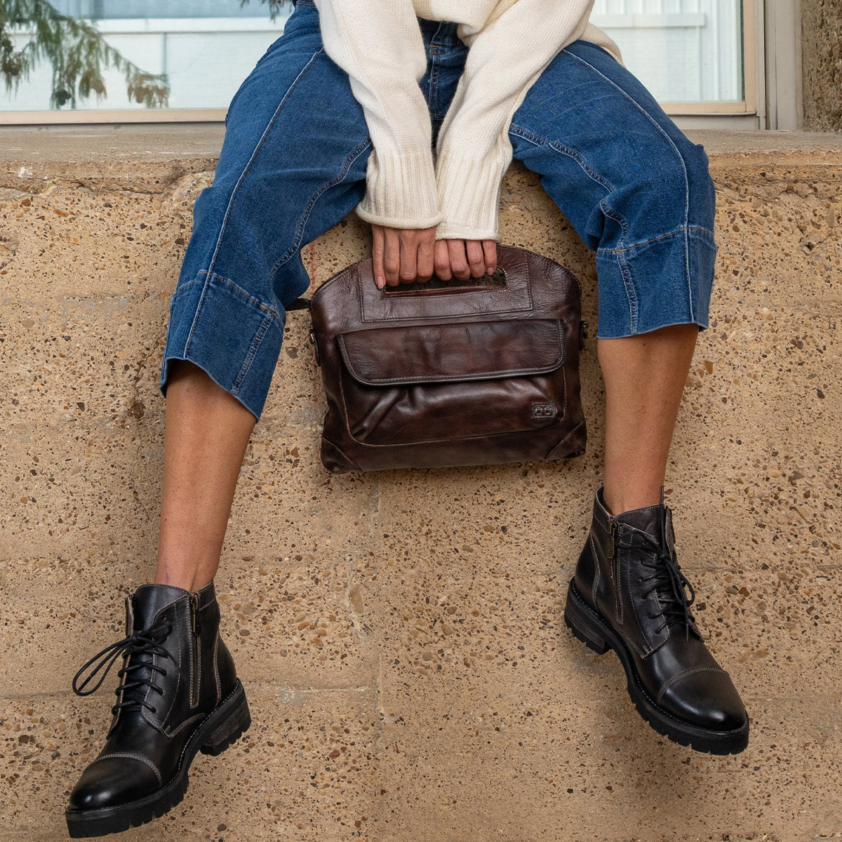 A person sitting on a ledge wears dark jeans and black lace-up ankle boots while holding a brown leather Bed|Stü Greenway zip-top handbag.
