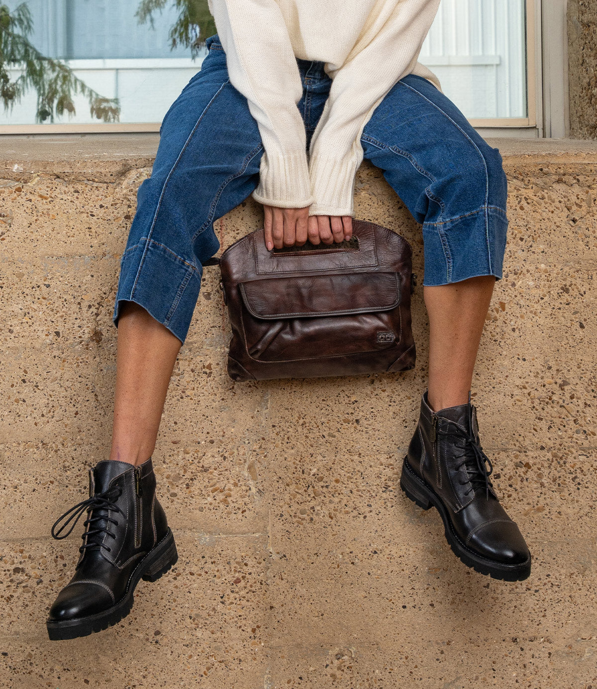A person sitting on a ledge wears dark jeans and black lace-up ankle boots while holding a brown leather Bed|Stü Greenway zip-top handbag.