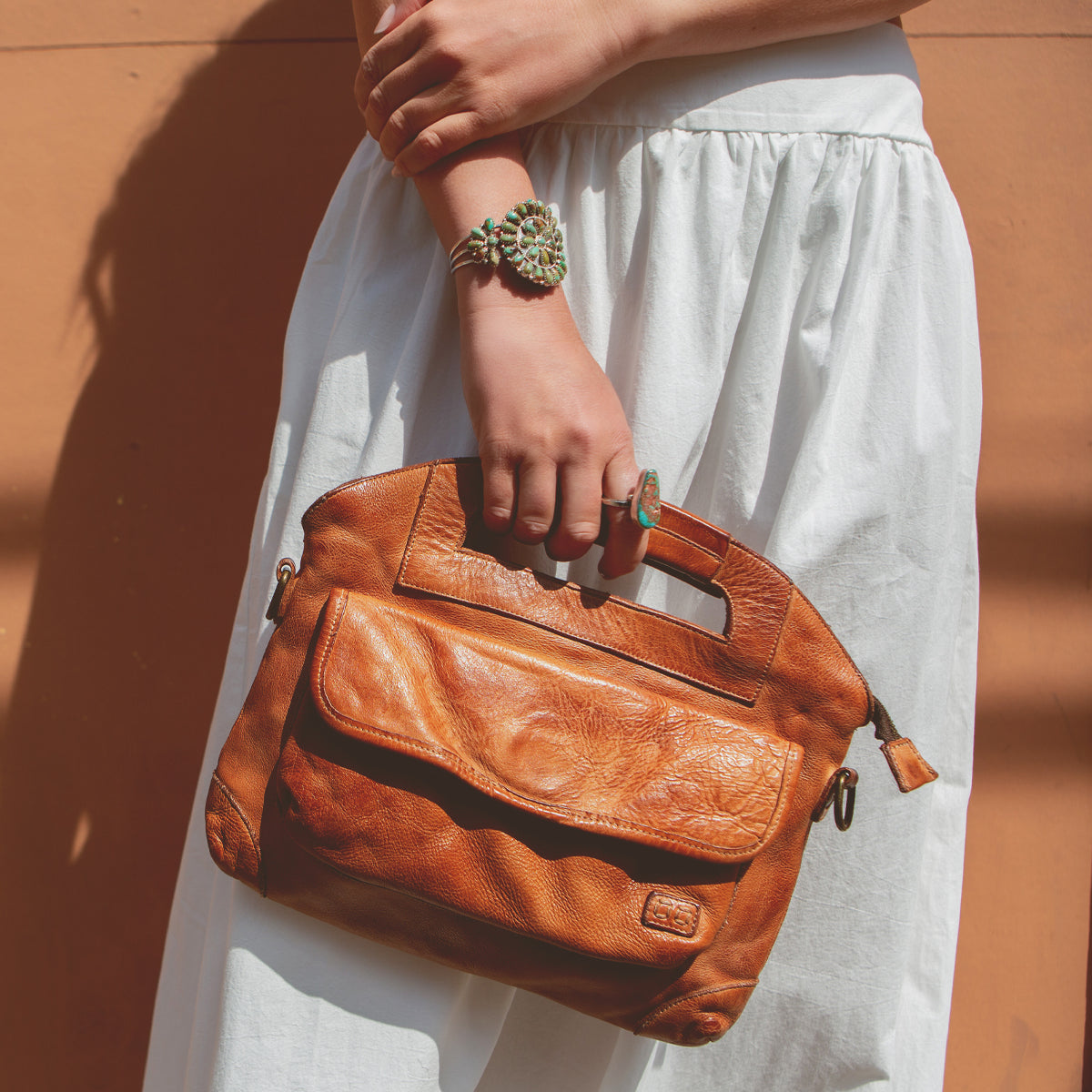 Woman holding a Greenway by Bed Stu leather handbag with a crossbody strap against a white skirt, against the backdrop of a terracotta wall.
