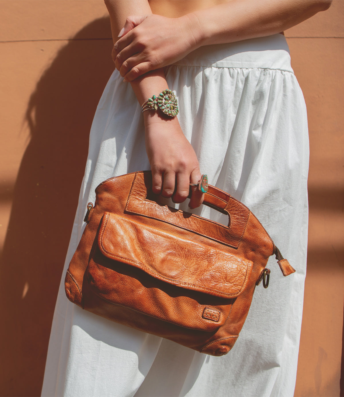 Woman holding a Greenway by Bed Stu leather handbag with a crossbody strap against a white skirt, against the backdrop of a terracotta wall.